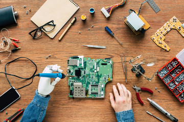 cropped image of electronic engineer with robotic hand fixing motherboard by soldering iron at table surrounded by tools - obrazy, fototapety, plakaty
