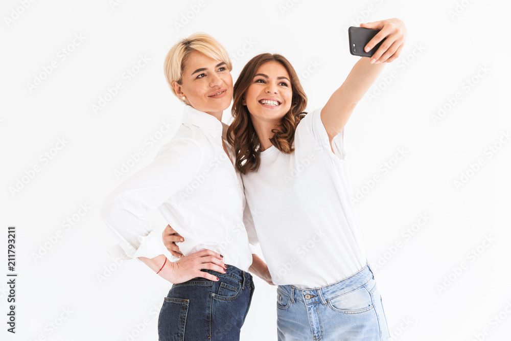 Poster Portrait of two joyous pretty women mother and daughter holding smartphone and taking selfie photo with smile, standing isolated over white background