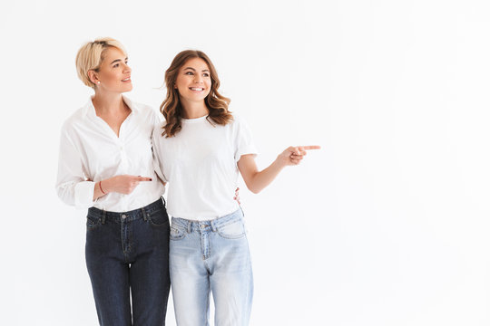 Picture of content two women mother 40s and daughter 16-18 pointing fingers aside at copyspace with smile, standing isolated over white background