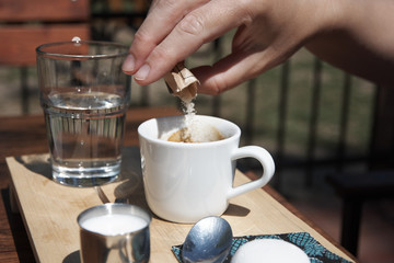 Young woman in a garden restaurant sprinkles sugar into the coffee. With a glass of water and a table.