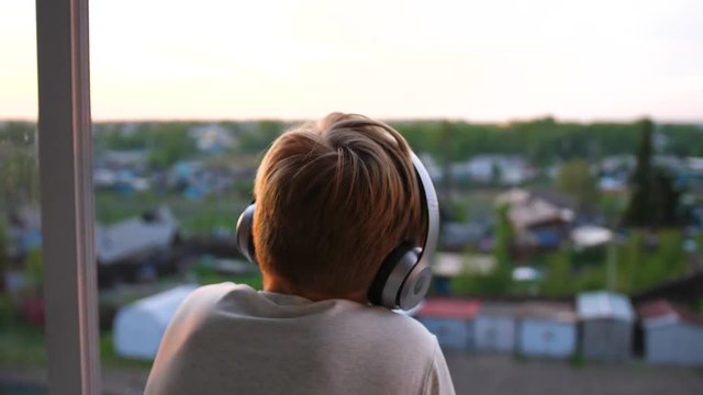A young man stands near the window and puts on headphones to listen to music. Blurred background with sunset, teen enjoying music in headphones.