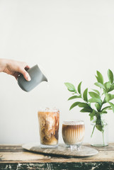 Iced coffee in tall glasses with milk pouring from pitcher by hand, white wall and green plant...