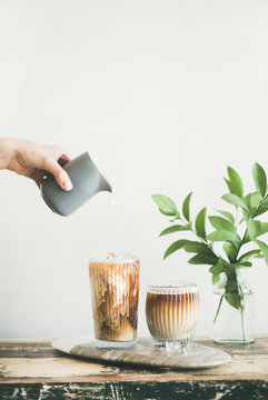 Iced coffee in tall glasses with milk pouring from pitcher by hand, white wall and green plant branches at background, copy space. Summer refreshing beverage ice coffee drink concept