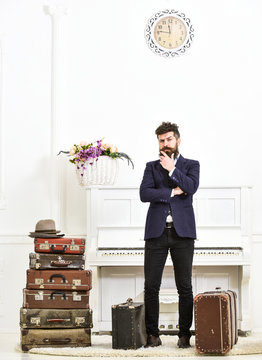 Macho elegant on thoughtful face standing near pile of vintage suitcase. Luggage and travelling concept. Man, traveller with beard and mustache with luggage, luxury white interior background.