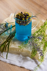 A glass brewer full of blue butterfly pea tea, with purple flowers and green branches next to it, on crumpled wrapping paper on a wooden table.