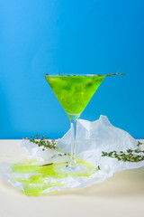 A green drink with ice cubes and green branches in a glass for mojito on crumpled wrapping paper on yellow table and blue background.