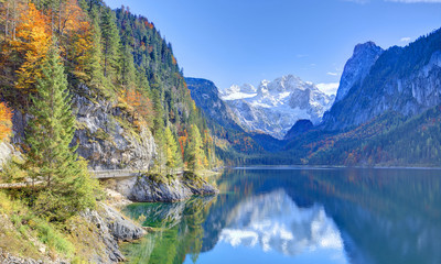 Naklejka na ściany i meble Autumn scenery of Lake Gosausee with snow-capped Dachstein Mountain in background & beautiful reflections on smooth water in Gosau, Austria ~ A dramatic scene of unspoilt nature of Alps on a sunny day