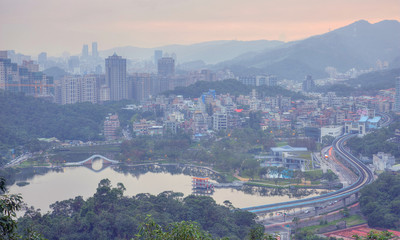 Aerial view of Da-Hu Community Park at dusk with an oriental arch bridge over the lake, nearby traffic trails on a busy street & residential blocks on the hillside in Neihu Dist., Taipei City, Taiwan