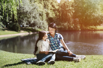 Pretty girls are sitting at the edge of small lake in park. They are looking at each other. Kid is holding opened book on her lap.