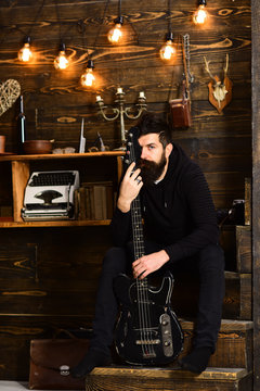 Connection through music. Man bearded musician enjoy evening with bass guitar, wooden background. Guy sits thoughtful dreamy in cozy warm atmosphere. Man with beard holds black electric guitar
