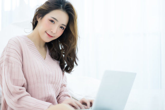Beautiful young Asian woman working on a laptop on a laptop sitting on bed in the house in the winter looking at camera. Woman lifestyle Concept