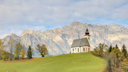 A church standing high on a grassy hilltop with a rocky mountain range in background~ Autumn scenery of St. Nikolaus Parish Church of Dienten Village at the foothill of Hochkoenig Mountains in Austria