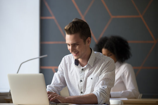Happy male millennial worker busy working at laptop in coworking space, satisfied employee chatting with friends or consulting client online at computer, man browsing web or surfing internet in office