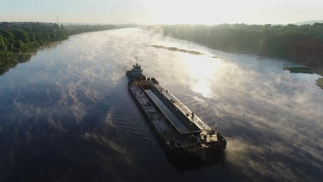 Aerial view. An old tug pushes a barge down a small river at dawn slowly. Thick fog beautifully spreads and reflected from the smooth surface of the water. Transport ship work on the river.