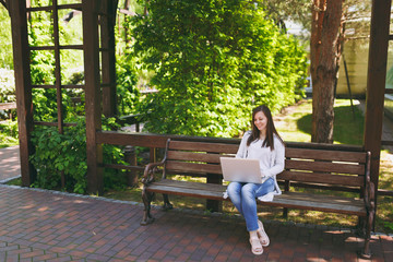 Obraz premium Young successful businesswoman in light casual clothes. Woman sitting on bench working on modern laptop pc computer in city park in street outdoors on nature. Mobile Office. Freelance business concept