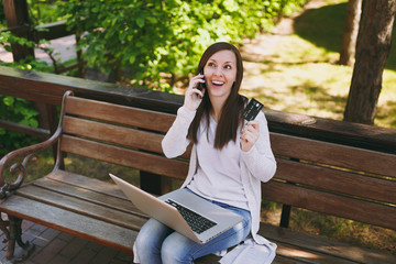 Fototapeta premium Female holding credit card, talking on mobile phone, having good pleasure conversation. Woman sitting on bench working on modern laptop in street outdoors. Mobile Office. Freelance business concept.