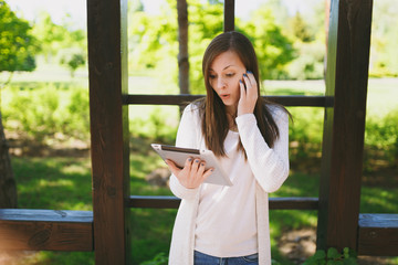 Portrait of sad upset shocked woman wearing light casual clothes. Pretty girl holding tablet pc computer, reading fake news in city park in street outdoors on spring green nature. Lifestyle concept.