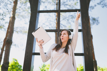 Naklejka premium Portrait of beautiful fun woman wearing light casual clothes. Pretty girl holding tablet pc computer, listening music in headphones in city park in street outdoors on spring nature. Lifestyle concept.