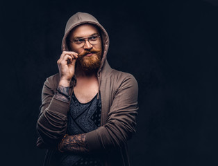 Pensive redhead hipster with full beard and glasses dressed in hoodie and t-shirt poses with hand on chin in a studio. Isolated on the dark background.