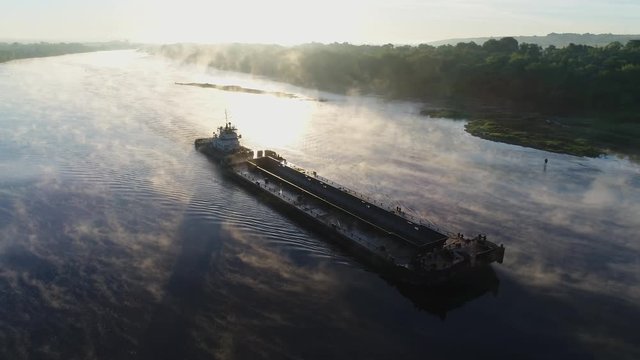 Aerial view. An old tug pushes a barge down a small river at dawn slowly. Thick fog beautifully spreads and reflected from the smooth surface of the water. Transport ship work on the river.