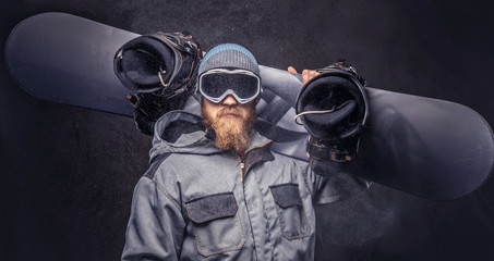 Brutal redhead snowboarder with a full beard in a winter hat and protective glasses dressed in a snowboarding coat posing with snowboard at a studio. Isolated on dark textured background.