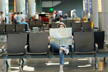Young traveler tourist woman covering with paper map, search route, waiting in lobby hall at international airport. Passenger traveling abroad on weekends getaway. Air travel, flight journey concept.