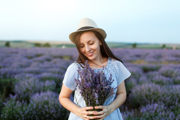 Young sensual beautiful woman in blue dress, hat on purple lavender flower blossom meadow field outdoors on summer nature background. Tender female near flowering bush with bouquet. Lifestyle concept.