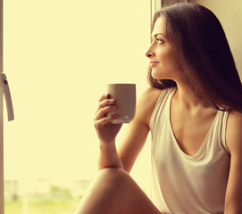 Beautiful smiling positive woman sitting near the window and looking up holding the cup of coffee in hand. Closeup toned color portrait