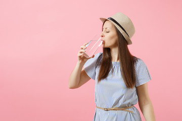Portrait of young beautiful woman in blue dress, hat holding and drinking clear fresh pure water from glass isolated on pink background. Healthy lifestyle, people, sincere emotions concept. Copy space