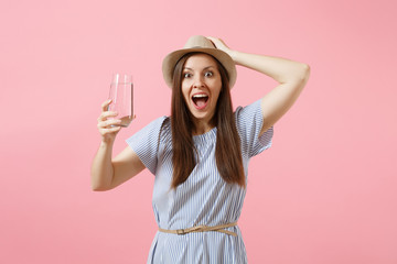 Portrait of young excited woman in blue dress, hat holding and drinking clear fresh pure water from glass isolated on pink background. Healthy lifestyle, people, sincere emotions concept. Copy space.