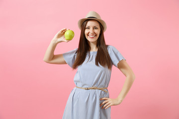 Portrait of young beautiful woman in blue dress, summer straw hat holding, eating green fresh apple fruit isolated on pink background. Healthy lifestyle, people, sincere emotions concept. Copy space.
