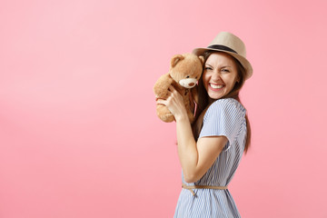 Portrait of young beautiful woman in blue dress, summer straw hat holding teddy bear plush toy isolated on pink background. People, sincere emotions, lifestyle concept. Advertising area. Copy space.