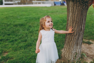 Naklejka premium Smiling little cute child baby girl in light dress stand and lean on tree on green grass in city park. Mother, little kid daughter. Mother's Day, love family, parenthood, childhood concept.