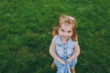 Fototapeta premium Smiling happy little cute child baby girl in denim dress looking up and have fun on green grass lawn in park. Mother, little kid daughter. Mother's Day, love family, parenthood, childhood concept.