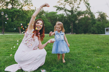 Naklejka premium Woman in light dress and little cute child baby girl play with colorful paper confetti, have fun in green park. Mother, little kid daughter. Mother's Day, love family, parenthood, childhood concept.