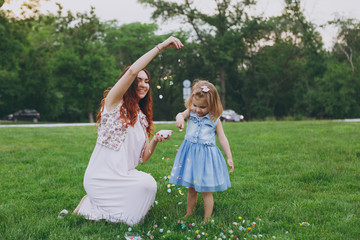 Obraz premium Smiling woman in light dress and little cute child baby girl play with colorful paper confetti in green park. Mother, little kid daughter. Mother's Day, love family, parenthood, childhood concept.