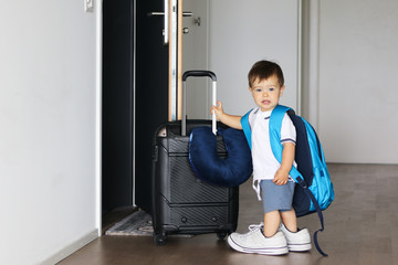 Cute little baby boy in fathers shoes and with big backpack holding suitcase staying near open door...