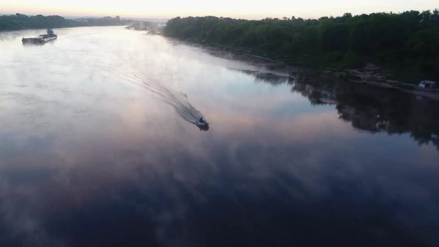 Aerial view. A small rubber fishing boat with a motor is moving quickly on a small river at dawn. The haze beautifully spreads over the smooth surface of the water. Sky and fog reflected in river.