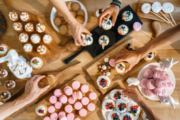 Many different confectionery products on a wooden table with peoples hands, top view. Gentle cake, pink macaroons, cookies, cake, marshmallows.