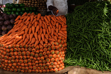 An indian man is selling fresh indian vegetables from his market stall. There are kakdi cuccumbers, melons, watermelons, carrots.
