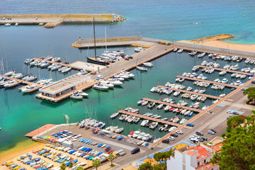 Fototapeta na wymiar Sea port with boat berths and concrete promenade. Private yachts and fishing boats are moored at the pier. Coast of Spanish beach resort Blanes in summertime. Costa Brava, Catalonia, Spain
