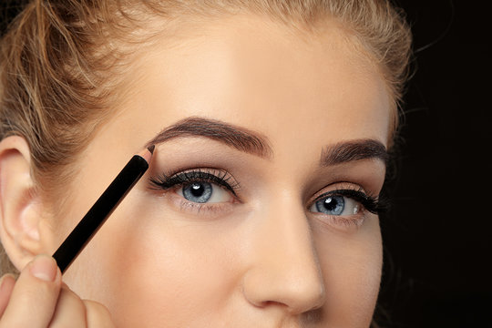 Young woman correcting shape of eyebrows on dark background, closeup