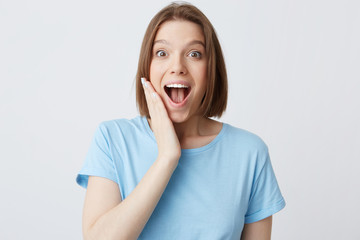 Closeup of happy excited attractive young woman in blue t shirt with opened mouth touching her cheek and feels surprised isolated over white background
