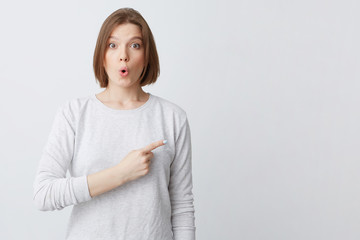 Obraz premium Amazed cute young woman in longsleeve making funny surprised face and pointing to the side with finger isolated over white background