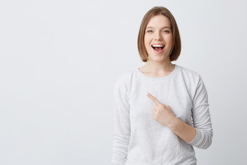 Happy excited young woman in longsleeve with opened mouth feels confident and pointing to the side isolated over white background