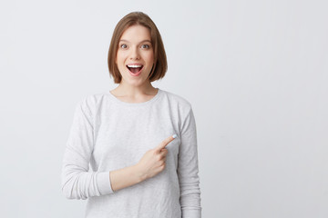 Obraz premium Surprised pretty young woman in longsleeve with opened mouth smiling and pointing to the side isolated over white background Feels happy and amazed