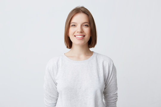 beautiful woman expresses happy emotions, has broad pleasant smile, wears white longsleeve and feels satisfied. Isolated over white background
