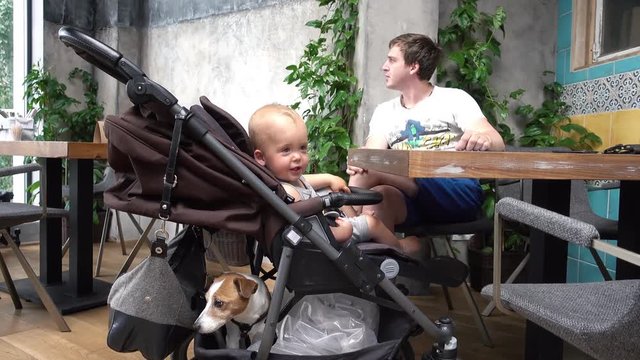 Baby boy sitting in stroller with dog and father in cafe. Happy joy caucasian little Child, kid toddler smiling in stroller. Sunday family dinner