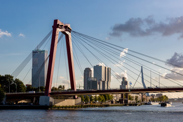 Orange cable bridge in Rotterdam with the cityscape of the Dutch city as a backdrop with the financial district and Erasmus bridge against a dramatic sunset sky with cumulus clouds over the skyline