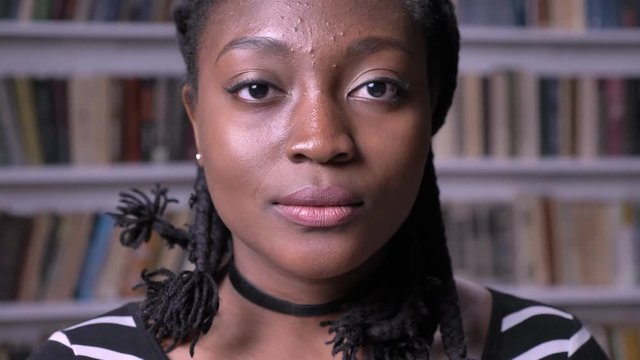 Portrait of young pretty african american woman looking at camera, thoughtful and concerned, standing in library with bookshelves